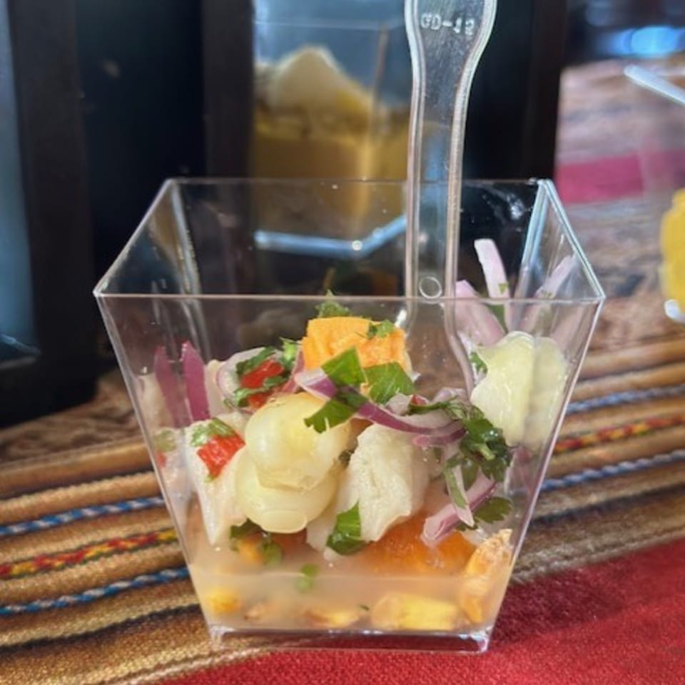 Peruvian ceviche in a clear square cup with a plastic spoon, on a colorful striped tablecloth.