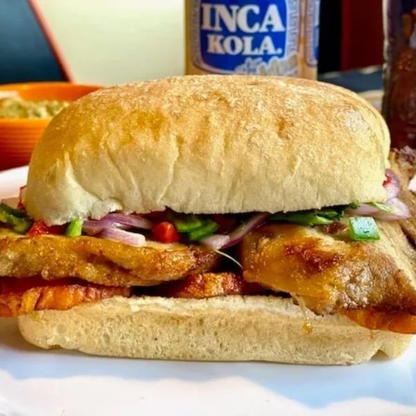 Close-up of a Peruvian chicken sandwich with red onions and green peppers, with an Inca Kola bottle in the background.