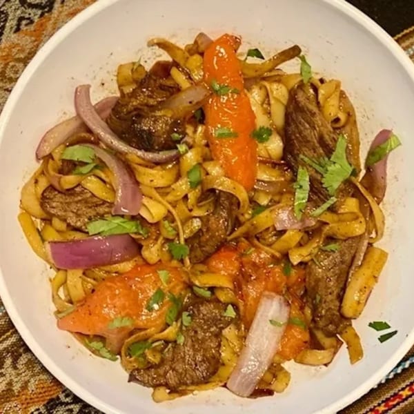 Peruvian Lomo Saltado with wide noodles, beef, red onions, tomatoes, and cilantro in a white bowl.